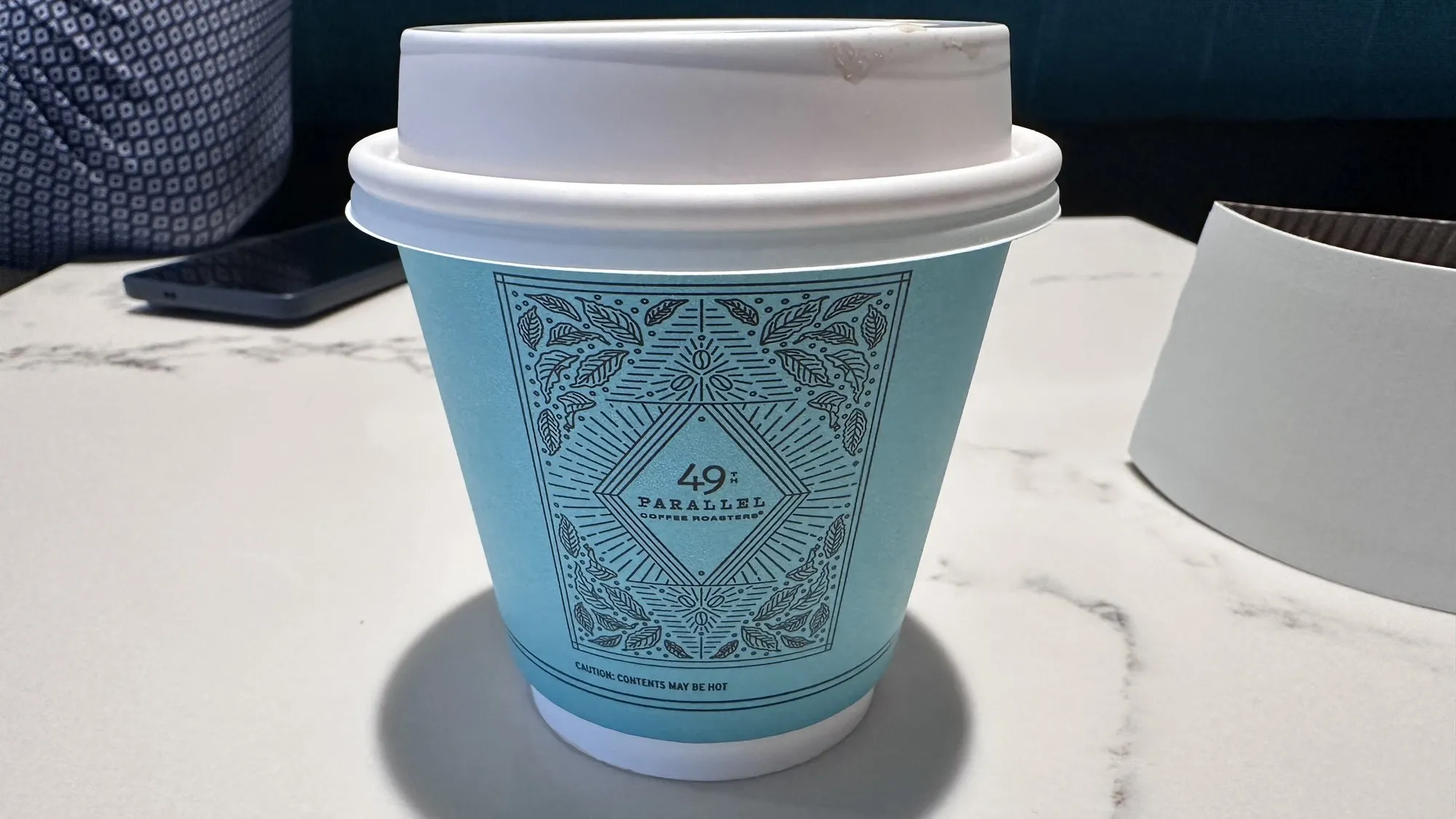 A teal 49th Parallel Coffee Roasters takeaway cup with an ornate leaf pattern, sitting on a white marble table.